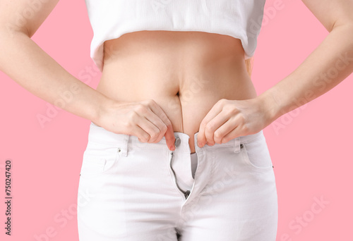 Young woman trying to button tight pants on pink background, closeup. Weight gain concept