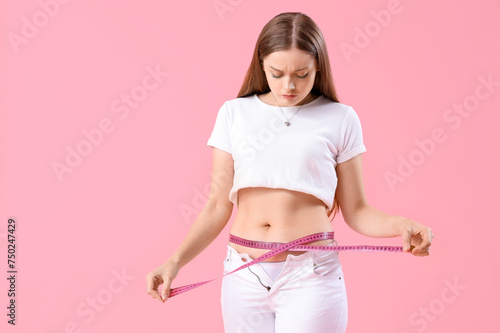 Young woman in tight pants measuring her belly on pink background. Weight gain concept © Pixel-Shot