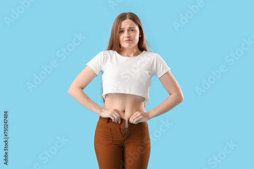 Upset young woman trying to button tight pants on blue background. Weight gain concept