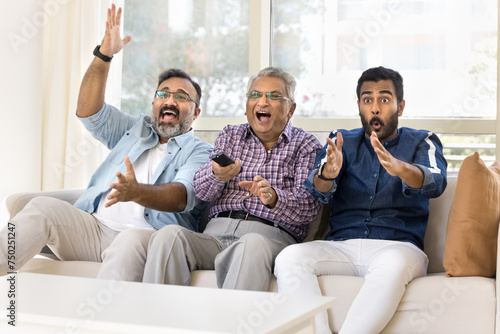 Excited happy adult Indian men of three family generations watching sport match on TV, celebrating team winning, laughing, shouting, waving hands, using remote control device, sitting on couch