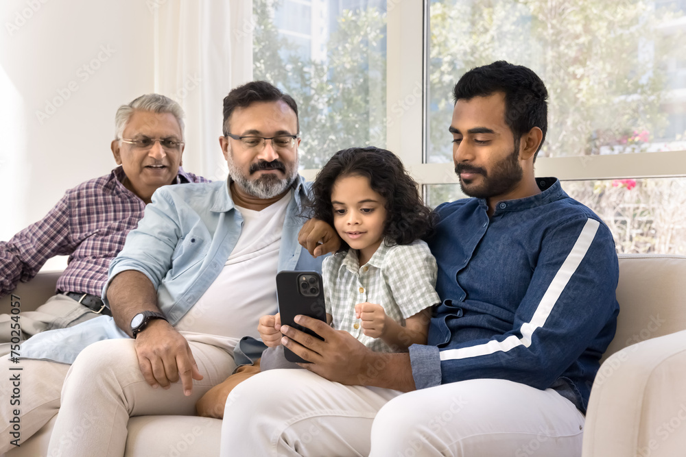 Focused Indian relatives of four different family generations using online application, media service for entertainment on cellphone at home, sitting on sofa together Focused Indian relatives of four different family generations using online application, media service for entertainment on cellphone at home, sitting on sofa together