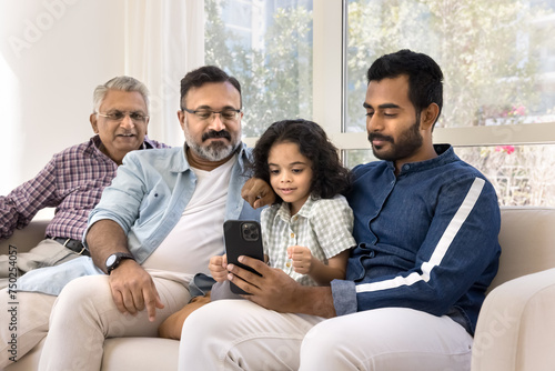 Focused Indian relatives of four different family generations using online application, media service for entertainment on cellphone at home, sitting on sofa together