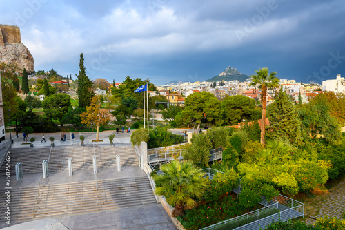 The tree lined Plaka neighborhood under the Acropolis Museum and ancient Acropolis Hill, with Mount Lycabettus covered in rain clouds in the distance, Athens Greece.