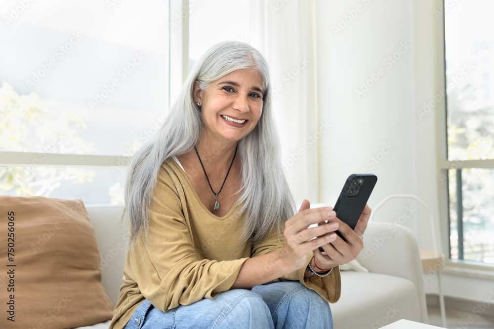 Cheerful positive grey haired senior Latin woman using online application, ecommerce service on mobile phone, posing for home portrait on white couch, looking at camera with toothy smile Cheerful positive grey haired senior Latin woman using online application, ecommerce service on mobile phone, posing for home portrait on white couch, looking at camera with toothy smile