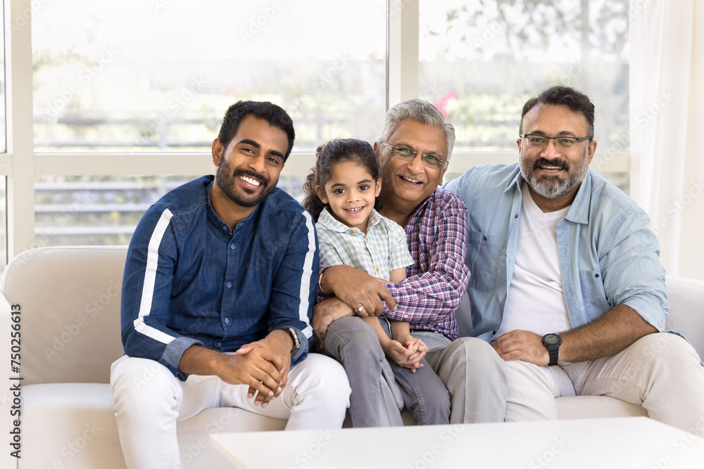 Happy Indian great grandpa holding little kid girl in arms on lap, sitting at mature son and young adult grandson on sofa, looking at camera. Four of family generations posing for home portrait Happy Indian great grandpa holding little kid girl in arms on lap, sitting at mature son and young adult grandson on sofa, looking at camera. Four of family generations posing for home portrait