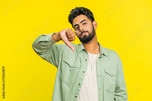 Dislike. Upset unhappy Indian man showing thumbs down sign gesture, expressing discontent, disapproval, dissatisfied negative feedback. Attractive bearded guy indoors isolated on yellow background