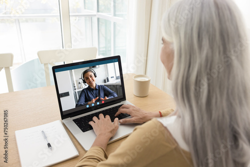 Positive young Indian woman in headset, online teacher, blogger, coach giving webinar, lecture, consultation, speaking online from laptop screen, training elderly student on Internet