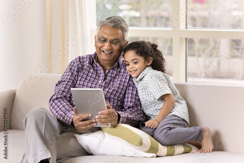 Happy Indian grandfather and little granddaughter girl using tablet for Internet communication, talking on family video call, sitting on home couch together, holding digital device, smiling