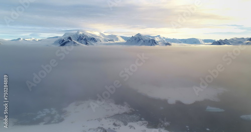 Fly over sunset clouds, mountain peak above sunrise fog cloudscape in Antarctica. Ocean aerial view snow covered surface of Antarctic continent. Wild nature background. Drone flight