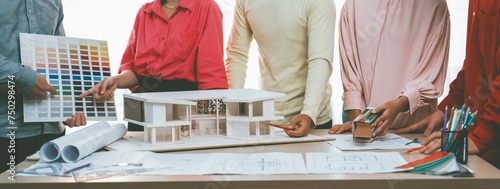 Cropped image of professional architect team brainstorm about house color selection while architect writes on blueprint document at table placed with blueprint and house model. Variegated.