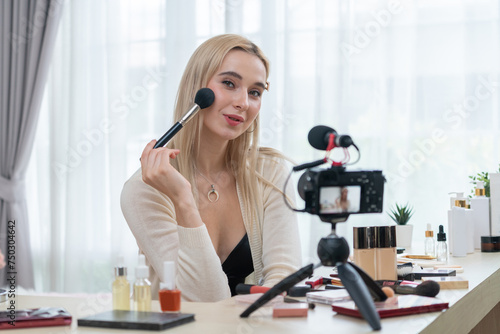 Young woman making beauty and cosmetic tutorial video content for social media. Beauty blogger smiles to camera while showing how to apply mascara to audience or followers. Blithe
