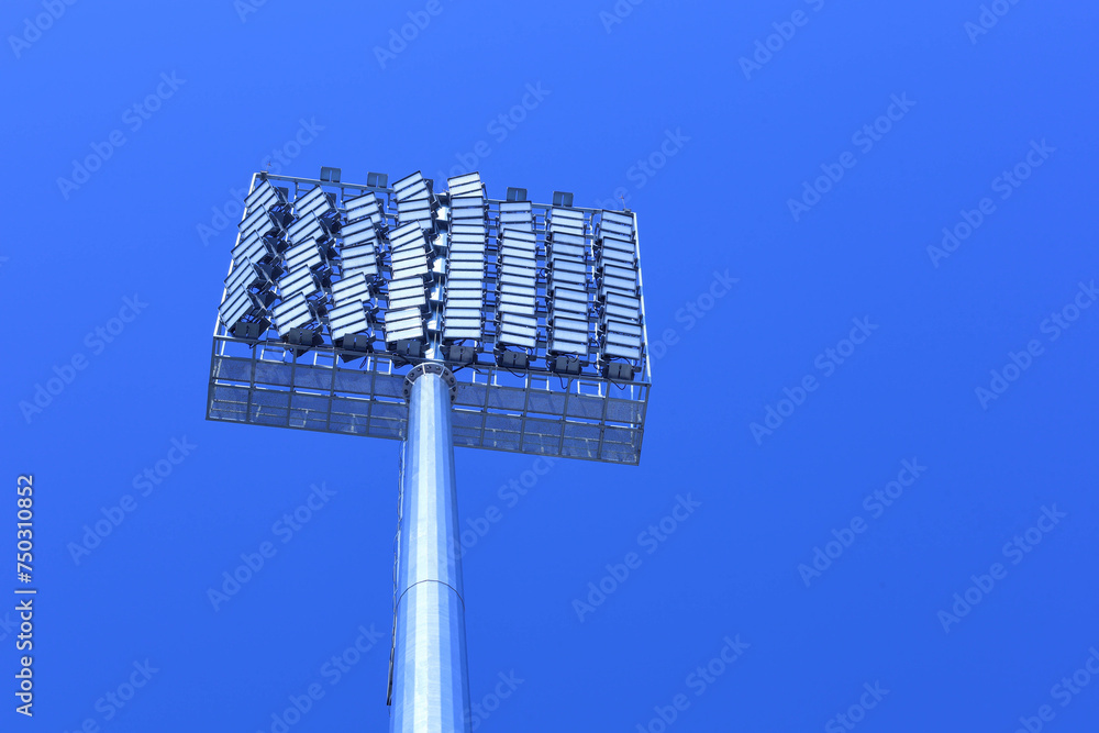 Closeup of stadium lights. Set of spotlight panels for outdoor stadium lighting on tall masts in bottom view on blue sky background with copy space with selective focus. Closeup of stadium lights. Set of spotlight panels for outdoor stadium lighting on tall masts in bottom view on blue sky background with copy space with selective focus.