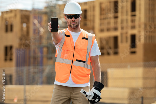 Builder in a hard hat working on a construction project at a site. Worker hold phone, builder show phone screen. Architect with mobile phone. Man worker in builders helmet on the building site.