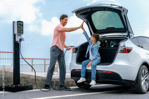 Family road trip vacation traveling by the sea with electric car, father and son high five after reach destination at EV charging station by the seashore. Eco-friendly car for environment. Perpetual © Summit Art Creations
