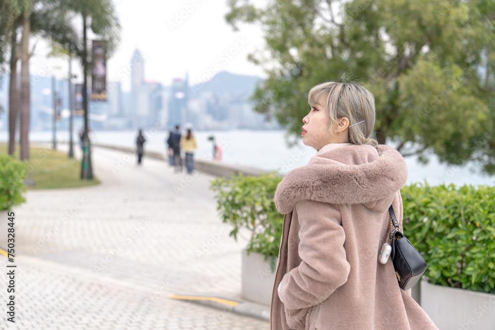 A young Chinese woman in her 20s dressed in winter walking in a park by the sea where you can see the skyscrapers of Kowloon, Hong Kong 香港九龍の高層ビルが見える海沿いの公園を散歩する冬の格好をした20代の若い中国人女性