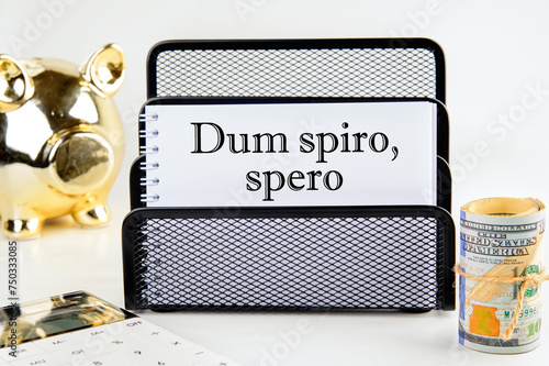 Dum Spiro Spero - latin phrase means While I Breath, I Hope. on a white notepad with money and a piggy bank in the background out of focus photo