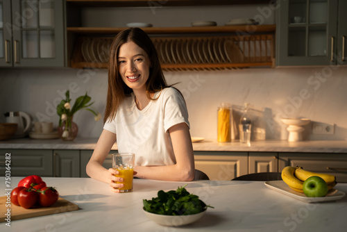 A young woman is seated at a kitchen counter  sipping from a glass of fresh juice  with a soft morning light illuminating her casual  relaxed pose. Fresh vegetables and fruits are laid out on the