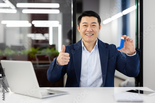 Portrait of a smiling young Asian man in a business suit sitting in the office, holding an asthma and allergy inhaler in his hand, and showing the camera a super gesture with his finger.