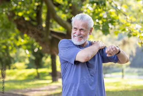 Fototapeta Naklejka Na Ścianę i Meble -  Senior gray-haired man sprained his ankle while walking in the park and exercising, standing outside and massaging his shoulder with his hand and feeling severe pain.