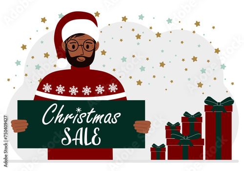 A man in a sweater and a red Santa hat holds a poster with the text about Christmas sale in his hand.