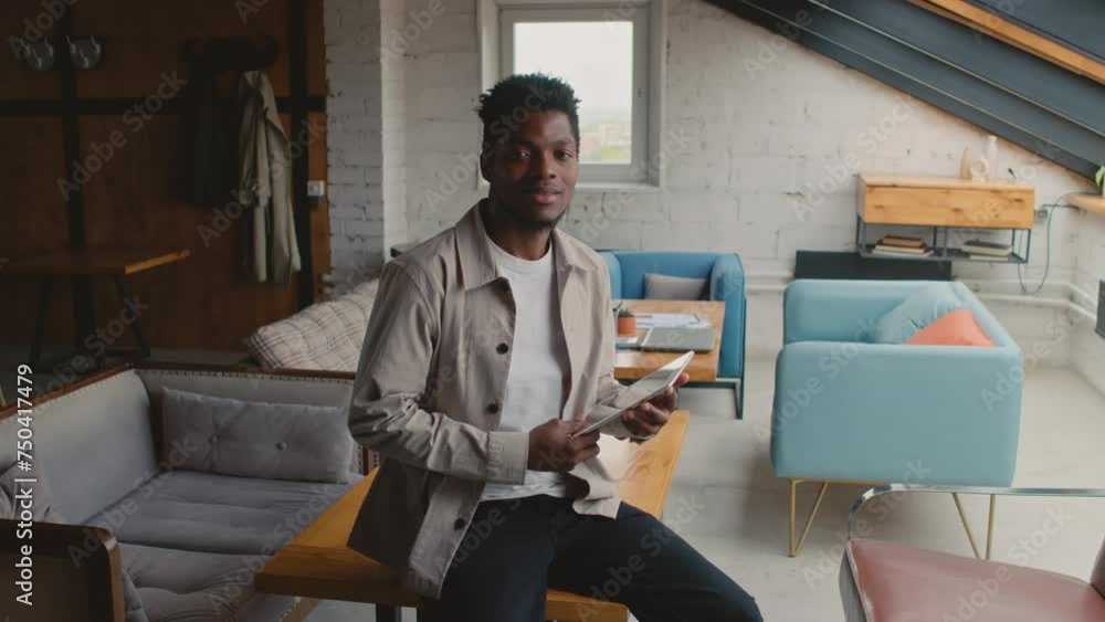 Medium portrait shot of young African American man in casual jeans and shirt sitting on table in coworking facility, holding tablet computer, smiling and looking at camera Medium portrait shot of young African American man in casual jeans and shirt sitting on table in coworking facility, holding tablet computer, smiling and looking at camera