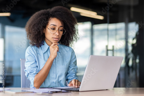 A serious young African American woman is sitting in the office, at desk in a blue shirt and is looking thoughtfully at the laptop screen.