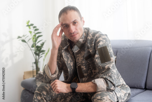 Portrait of calm serious Caucasian military man wearing camouflage uniform and cap sitting on a sofa, having depressed facial expression.