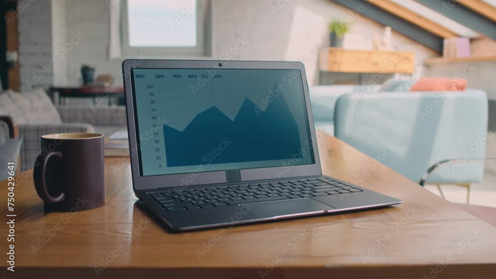 Close-up no people shot of laptop computer with financial data and graph on screen, and coffee mug on table in empty loft-style coworking office Close-up no people shot of laptop computer with financial data and graph on screen, and coffee mug on table in empty loft-style coworking office