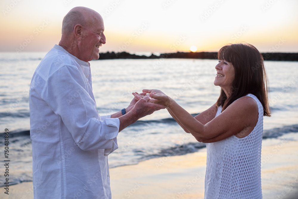Smiling bonding senior couple standing at the beach holding hands looking in the eyes enjoying vacation and retirement expressing love and tenderness