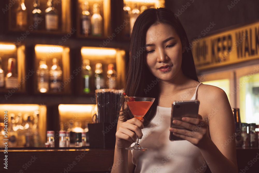 Asian woman used to chat with her friends on cellphone while drinking whiskey at the bar.