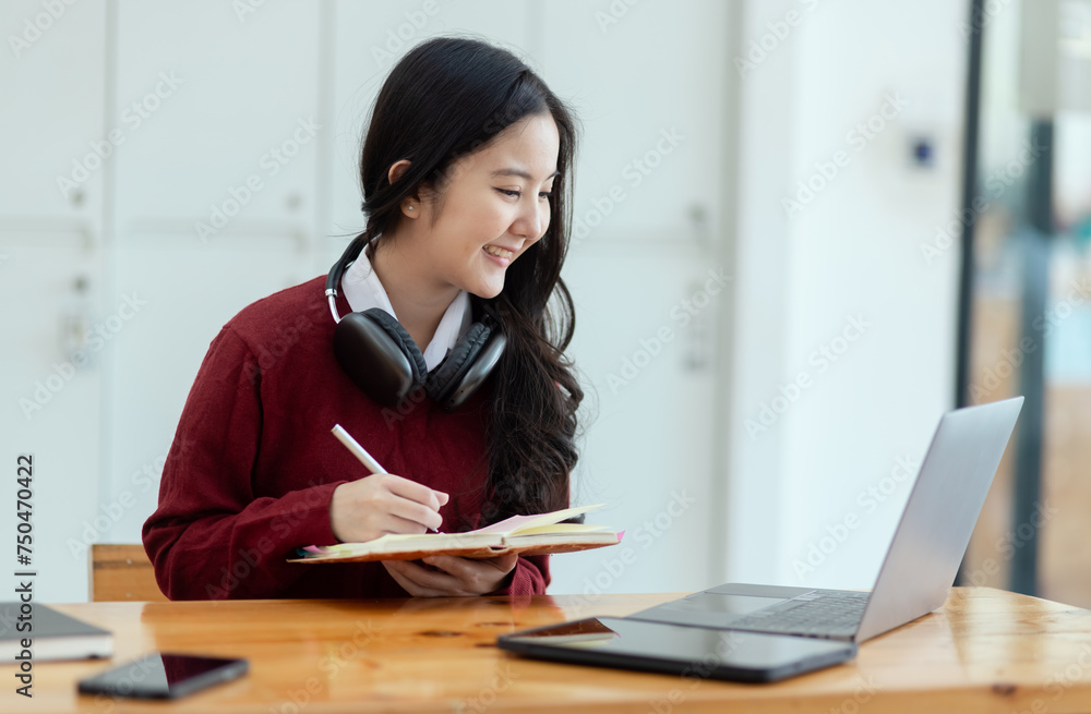 Happy young asian woman wearing earphones and using laptop computer at desk in office, Female student with stack of books and laptop. Happy young asian woman wearing earphones and using laptop computer at desk in office, Female student with stack of books and laptop.