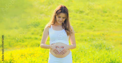 Happy young pregnant woman makes heart sign on her belly with hands in summer park