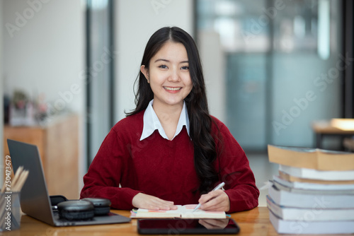 Happy young asian woman wearing earphones and using laptop computer at desk in office, Female student with stack of books and laptop.