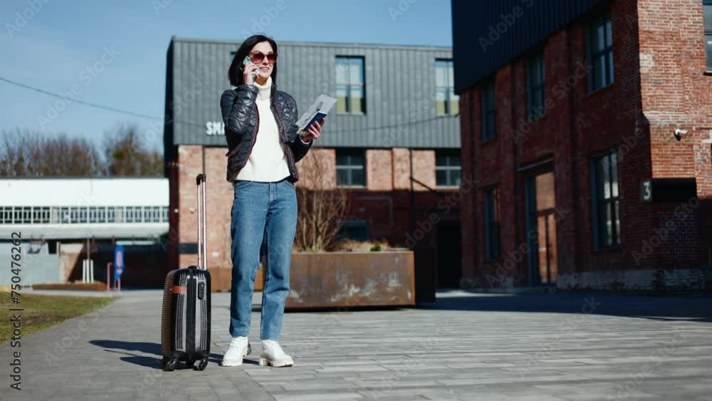 Relaxed lady in casual outfit standing next to small suitcase and answering smartphone call on buildings background. Positive female waiting for transport to train station for going on business trip. Relaxed lady in casual outfit standing next to small suitcase and answering smartphone call on buildings background. Positive female waiting for transport to train station for going on business trip.