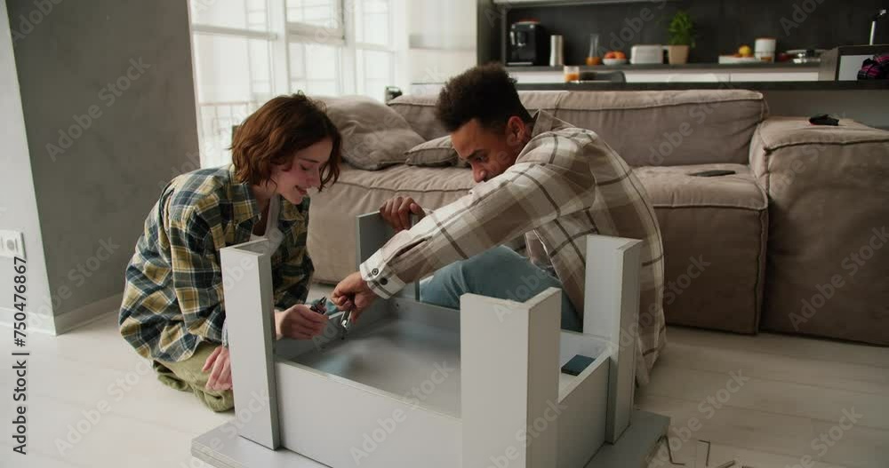 Confident and happy young man with Black skin color in a beige shirt is repairing and assembling a table together with his young adult girlfriend with a bob hairstyle in a plaid shirt sitting on the Confident and happy young man with Black skin color in a beige shirt is repairing and assembling a table together with his young adult girlfriend with a bob hairstyle in a plaid shirt sitting on the