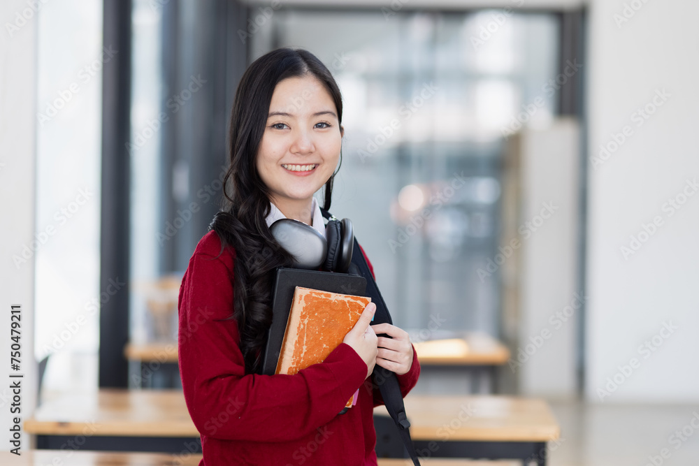 Fototapeta premium Happy young asian woman wearing earphones and holding a book in a classroom at high school.