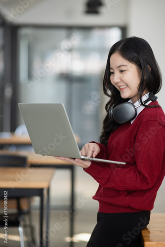 Happy young asian woman wearing earphones and using laptop computer at desk in office.