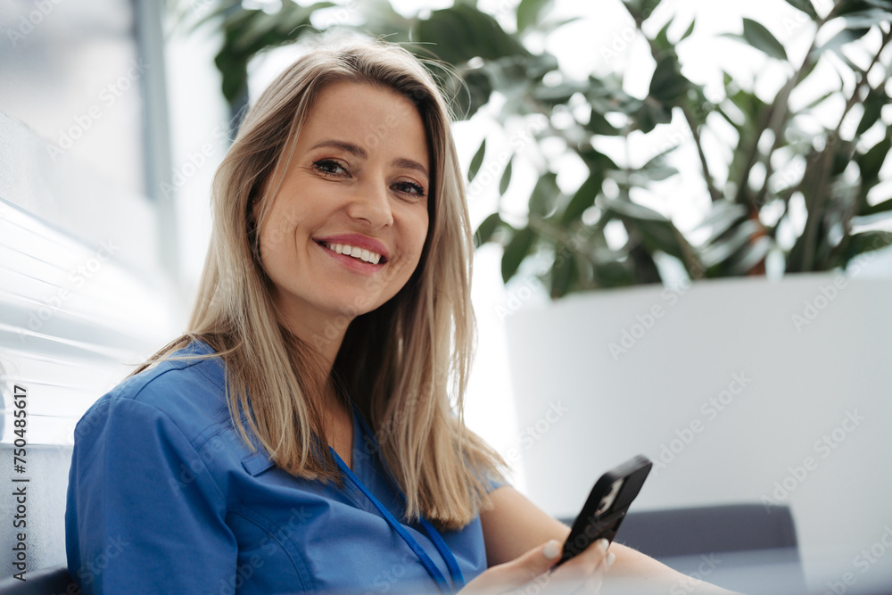 Fototapeta premium Beautiful female doctor taking a break during her work shift at hospital, scrolling on smartphone.