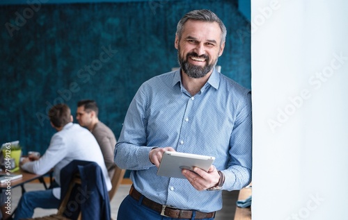Smiling elegant mid aged business man wearing suit standing in office holding digital tablet