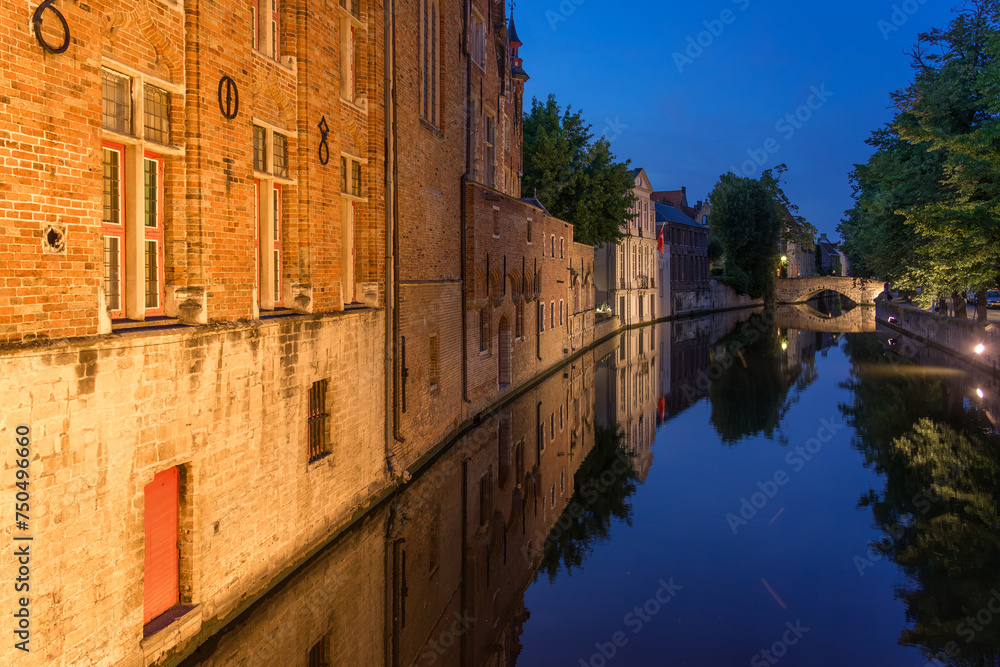 Historic buildings reflected on the canal in the old town of the beautiful city of Brugge in Belgium with the Meestrat bridge in the background at night.