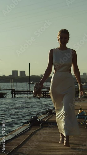 Stylish and beautiful woman in trendy attire, a long white dress, and sandals, strolling along the pier by the water.