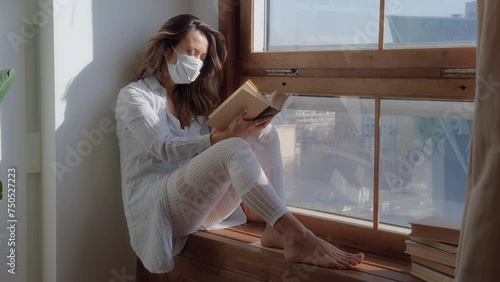 depressed native American young woman with medical mask sits on window sill at home alone reading, loses heart and throws book, feels sad due to sick and loneliness photo