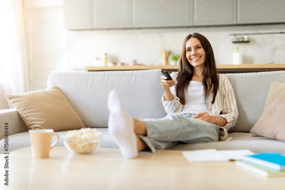 Smiling young woman lounging on couch with feet up, holding TV remote