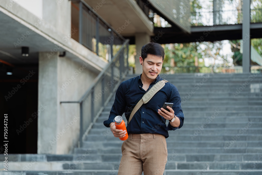 Smiling asian businessman hold reusable eco-friendly ecological cup and using mobile while on the way home at park Smiling asian businessman hold reusable eco-friendly ecological cup and using mobile while on the way home at park