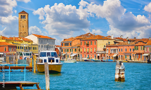 Panoramic view of Murano island, Venice, Veneto, Italy. At bell tower brick building Church Santa Maria e San Donato. Wooden dock with boat on the water and scenic sky summer clouds.