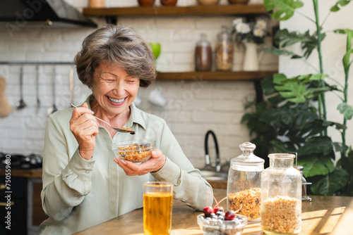 Cheerful senior grandmother eating cereals for breakfast mixing with berries drinking apple juice at home kitchen. Healthy dieting concept. Active seniors