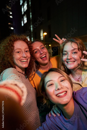 Quadro su tela Four happy smiling young female friends taking a vertical selfie looking at camera together at night outdoors
