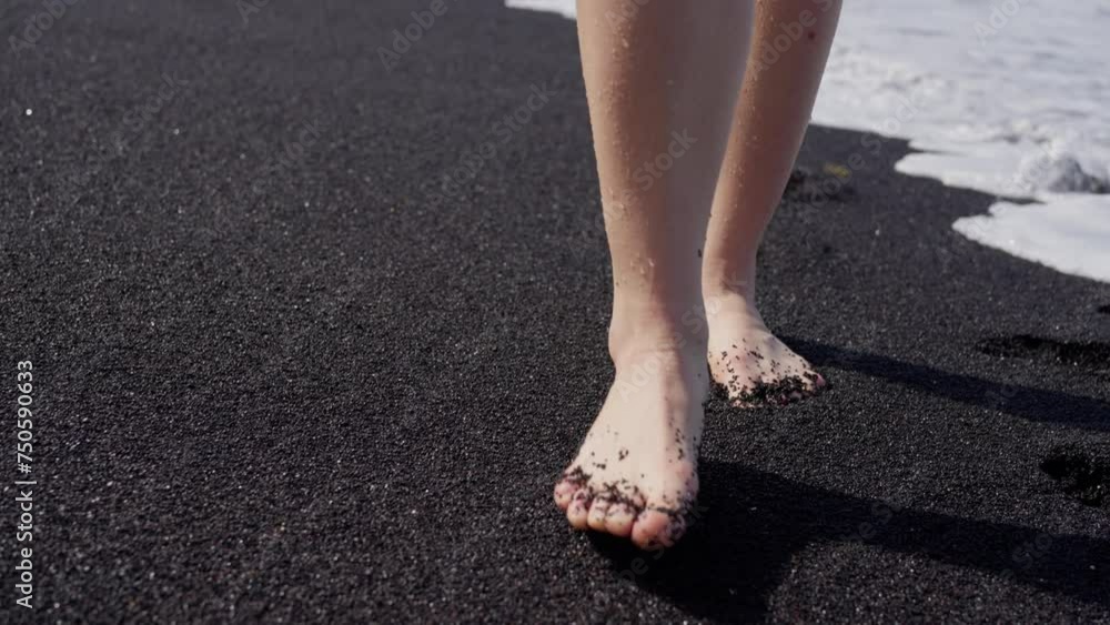 A woman, walks barefoot on the black sandy beach, the water gently caressing her feet as she moves gracefully along the shore. Girl is strolling on black sand barefoot, by the water. Slow motion A woman, walks barefoot on the black sandy beach, the water gently caressing her feet as she moves gracefully along the shore. Girl is strolling on black sand barefoot, by the water. Slow motion