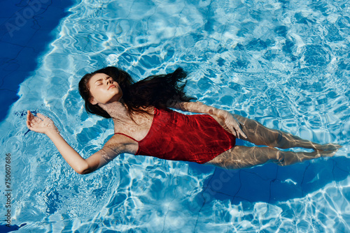 Young woman in the pool in red swimsuit with a beautiful smile lying on the water and swimming in the blue pool in the sun, the concept of relaxing on vacation.