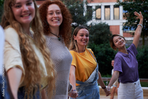 Four happy and friendly young women holding hands posing in a row looking at the camera, smiling and having fun outdoors. Group of female friends enjoying together in a city park.